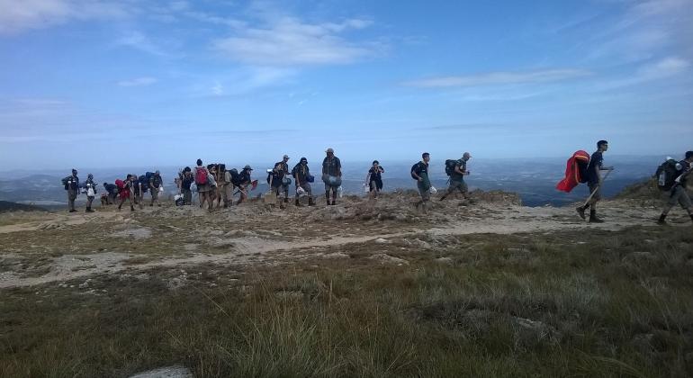 Cerca de 16 escoteiros fazem cainhada em local verde e alto em dia de céu azul. 