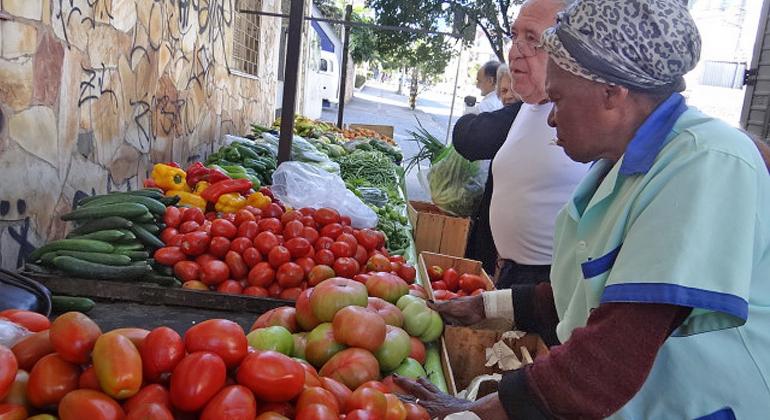 Mulher vende tomates a homem em banca durante o dia.