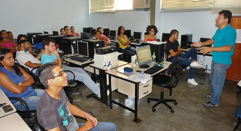 Cerca de nove jovens sentados em sala com mesas e computadores e Instrutor de pé. Foto ilustrativa