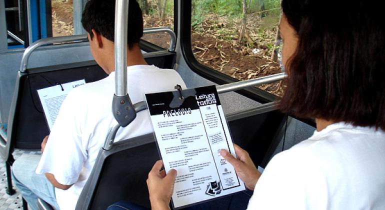 Dois passageiros de ônibus, sentados, lêem textos do Projeto Leitura para Todos, afixados na cadeira em frente ao passageiro. 