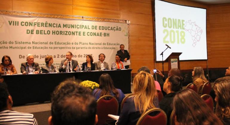 A foto mostra um auditório com algumas pessoas olhando para um palco onde está uma mesa com sete pessoas e uma mulher de pé. Ao lado , na parede, vê-se uma tela de projeção, onde se lê Conae 2018. Há uma faixa atrás da mesa onde se lê VIII Conferência Municipal de Educação de Belo Horizonte e Conae BH. 