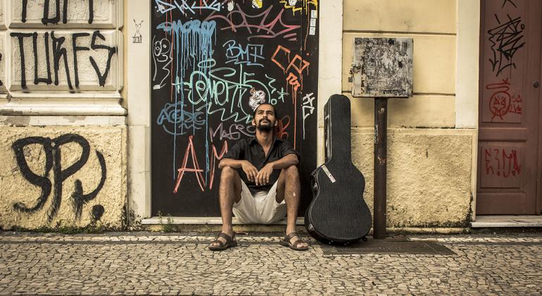Músico Moisés Pescador sentado ao lado de uma caixa de violão, na entrada de uma casa antiga, com porta e muros pixados e tendo a frente, uma calçada portuguesa.