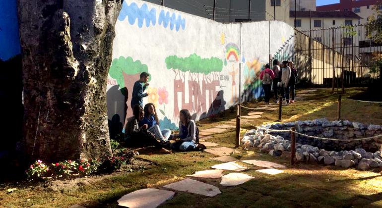 Cerca de oito estudantes, quatro deles sentados junta à árvore e outros quatro perto da grade, aproveitam o dia próximo a nascente revitalizada pela Escola Municipal Santos Dumont.