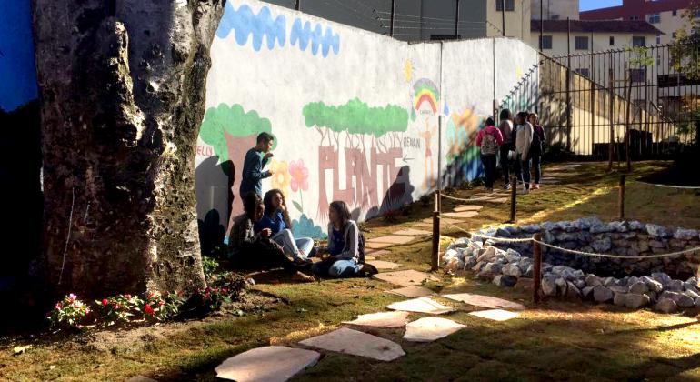 Escola Municipal Santos Dumont recupera nascente em seu terreno; ação faz parte de projeto do Comitê de Bacia Hidrográfica do Rio das Velhas. Foto: Smed/PBH Cerca de oito estudantes, quatro deles sentados junta à árvore e outros quatro perto da grade, aproveitam o dia próximo a nascente revitalizada pela Escola Municipal Santos Dumont.