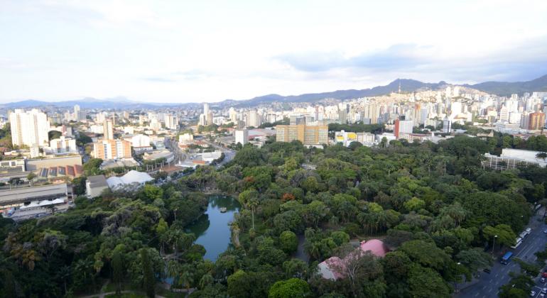 Foto: Amira Hissa/PBH O verde do Parque Municipal Américo Renneé Giannetti com Avenida Andradas à frente; prédios de Belo Horizonte ao fundo; mais ao fundo, montanhas e céu claro.