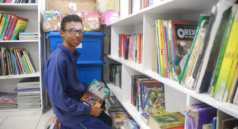 Aluno de escola municipal, ajoelhado, escolhendo livros em uma prateleira.
