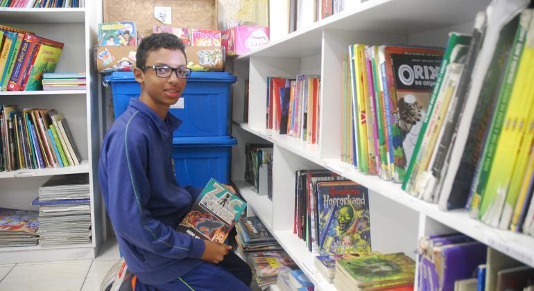 Escola da região Nordeste sensibiliza alunos para o universo da leitura e da escrita com projetos em biblioteca. Foto: Mara Damasceno/PBH Aluno de escola municipal, ajoelhado, escolhendo livros em uma prateleira.