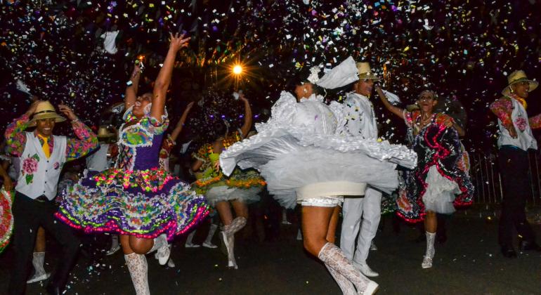 Quadrilha junina com roupas coloridas e mais de cinco membros dança e joga confetes à noite. 