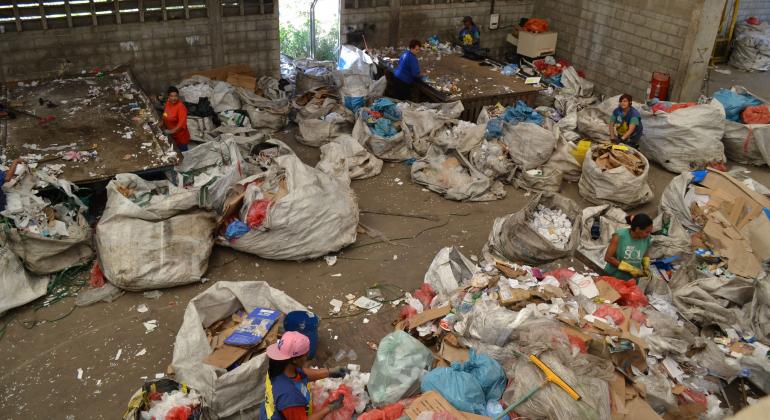 Galpão de reciclagem de lixo, com muitos sacos de lixo e uma mulher cercada de copos plásticos.