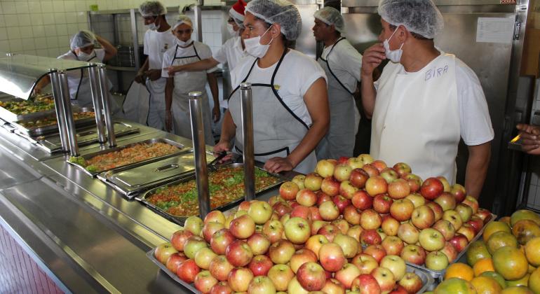 Sete funcionários de restaurante popular na mesa de almoço para servir comida; vê-se maçãs, laranjas e cardápio de almoço.