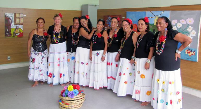Nove integrantes do Grupo de Cultura Popular Rosas do São Bernardo, com saias coloridas, rosa o cabelo e colares.