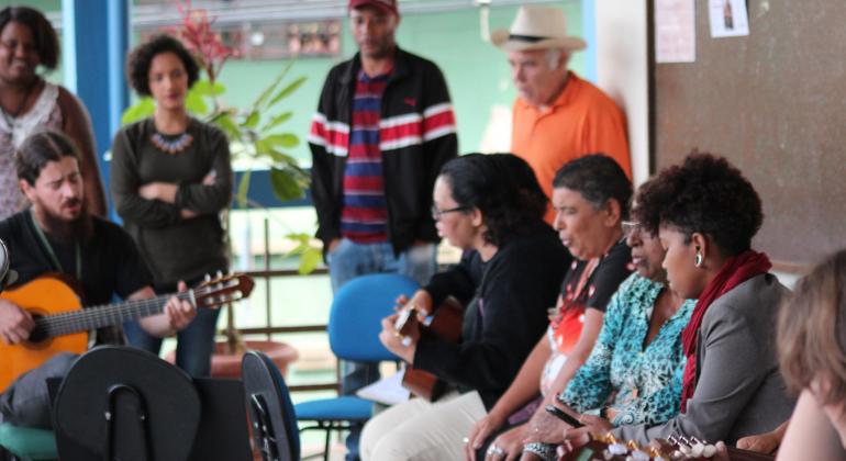 Dois homens com tocam violôes e sete pessoas, das quais quatro estão em pé, assistem ou cantam junto, atividade do projeto Varanda Musical, do Centro Cultural Alto Vera Cruz.