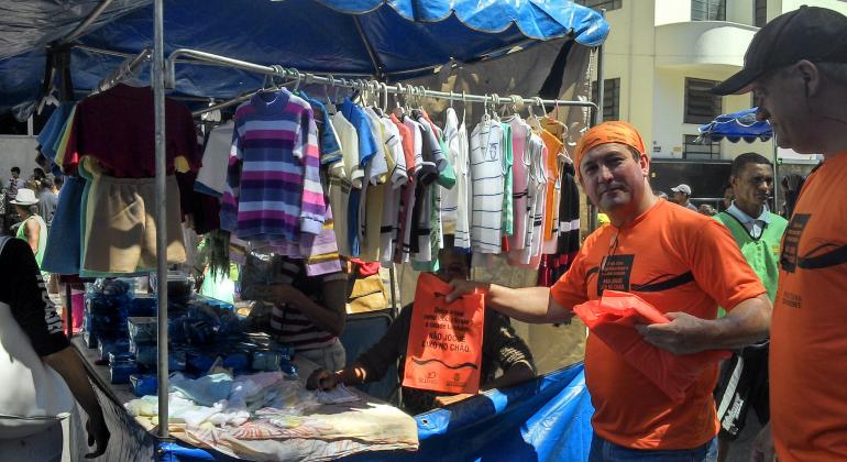 Sensibilização de feirantes e frequentadores contribui para a manutenção da limpeza da Feira da Afonso Pena. Foto: Divulgação SLU/PBH Membro da campanha de conscientização para limpeza com blusa, lenço e saquinho com mensagens educativas.