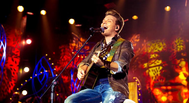 Foto: Divulgação O cantor sertanejo Michel Teló toca violão sentado em banco alto em show musical.