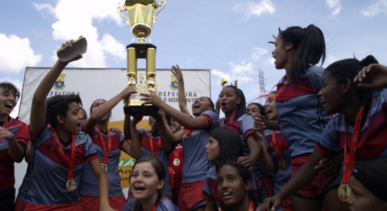Cerca de doze integrantes da equipe feminina de futebol Tupinambás, campeâ de 2016, erguem a taça e comemoram.