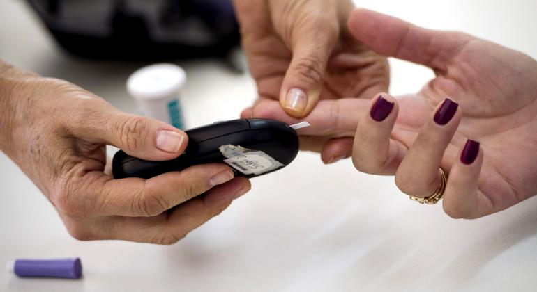 Mão femininaestá estendida com o dedo indicador em riste ao lado de mãos masculinas com o aparelho de medir glicemia com tira de exame. Ao fundo, desfocado, um tubinho de tiras. 