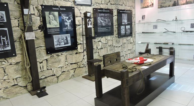 Sala do Museu da Inconfidência com mesa histórica e fotos e documentos expostos em parede de pedra e estante de vidro. 