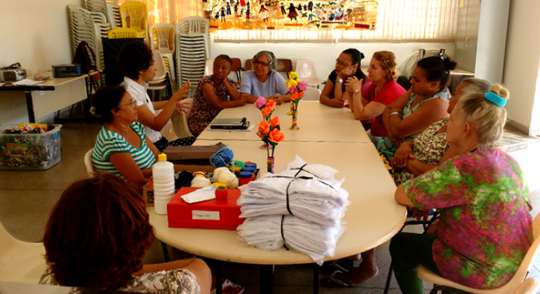 Nove mulheres reunidas em torno de mesa com panos, linhas e cadernos.