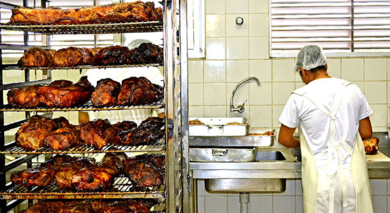 Confira os cardápios diários dos restaurantes populares de 28 de agosto a 1 de setembro. Foto: Denis Dias/PBH Mais de vinte frangos assados em prateleiras de metal na cozinha de restaurante popular; à direita, três formas retangulares de metal com frangos assados; mais à direita, funcionário corta frango assado em tábua de cozinha.