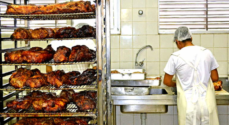 Mais de vinte frangos assados em prateleiras de metal na cozinha de restaurante popular; à direita, três formas retangulares de metal com frangos assados; mais à direita, funcionário corta frango assado em tábua de cozinha.