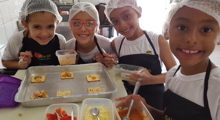 Quatro meninas vestidas com aventais e tocas de cozinha posicionadas em volta de uma mesa sorrindo e segurando vasilhas com comida. Em cima da mesa um tabuleiro com biscoitos dentro