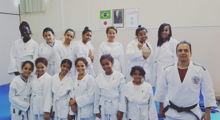 Treze garotas vestidas com uniforme branco de judô, posicionadas em duas fileiras, na fileira de trás as sete meninas em pé sorrindo e na fileira da frente seis meninas ajoelhadas juntamente com o professor ajoelhas. 