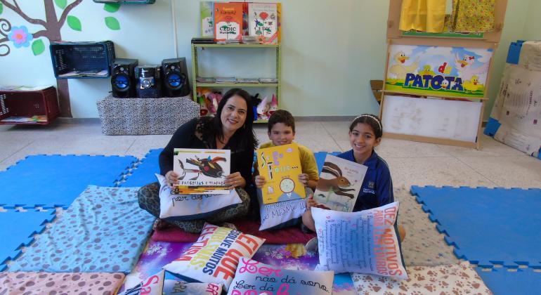 Professora e duas crianças sentados em espaço literário de escola e seguram almofadas escritas