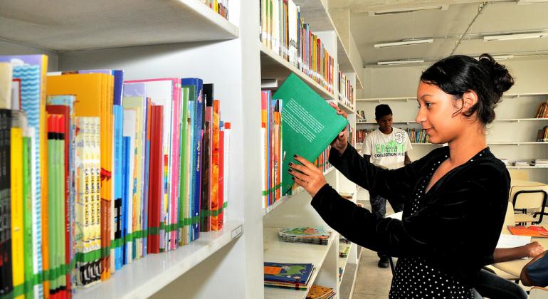 Prefeitura sorteia bolsas parciais e integrais Menina mexe em livro em estante de biblioteca