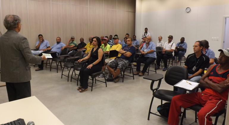 Adultos sentados em sala, assistindo palestra de homem.