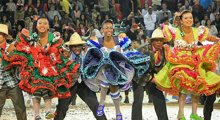 Três mulheres e dois homens vestidos com roupas de festejos juninos dançam e se apresentam ao público. 