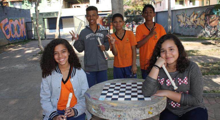 Cinco crianças, duas garotas e três garotos, três delas com flautas, próximos à mesa de xadrez de jardim, em lugar aberto.
