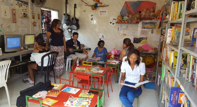 Sete pessoas utilizam biblioteca comunitária, realizando atividades diversas como ler, tocar violão e utilizar o computador.