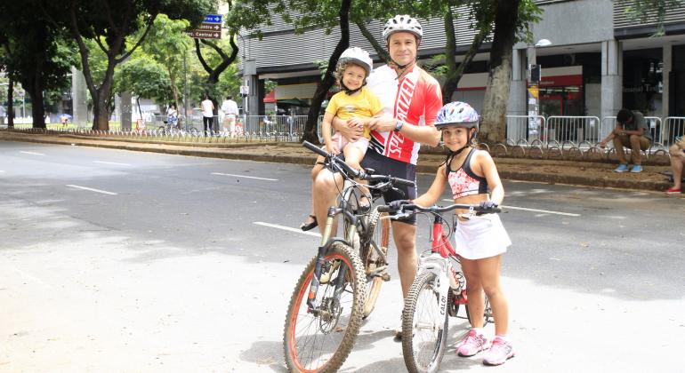 Homem de bicileta e capacete com filho no colo de capacete e garota ao lado, com bicileta e capacete.