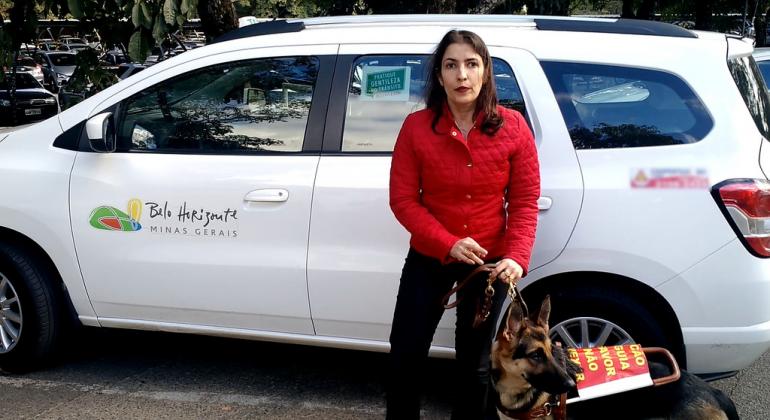 Mulher deficiente visual em frente à táxi com cão-guia de raça pastor alemão.