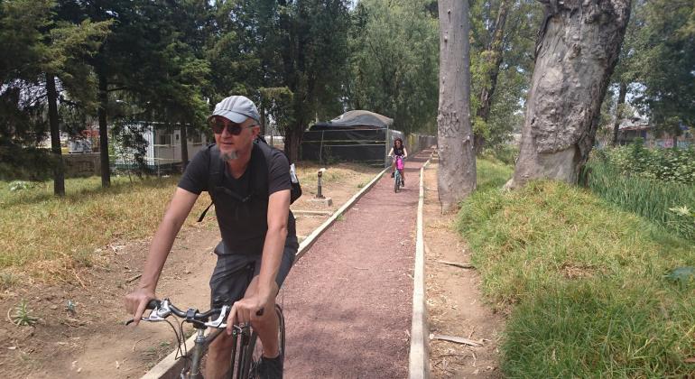 Homem anda de biclicleta em ciclovia em local arborizado. Ao fundo, outra pessoa de bicicleta.