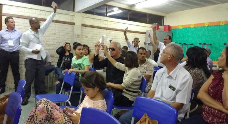 Cerca de cinco pessoas levantam a mão segurando cartões brancos em sala com mais de vinte pessoas, a maioria delas sentada.