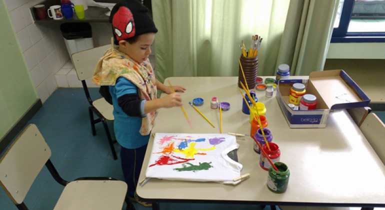 Criança com toca do Homem aranha pinta camiseta branca em mesa; à sua frente, vários potes de tinta.