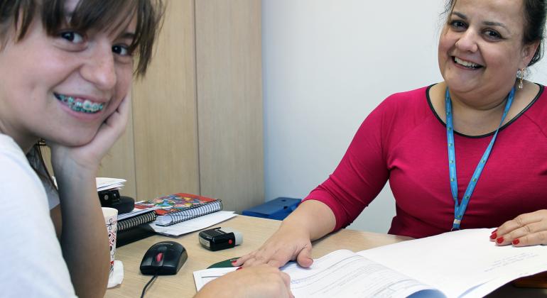 Com novo sistema de inscrições, programa emprega mais de mil estagiários na administração direta e é oportunidade de aprendizado. Foto: Planejamento/PBH Estagiária sentada em frente a mesa de escritório; à frente, servidora da Prefeitura.