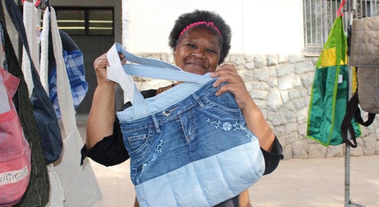 Mulher mostra bolsa artesanal feita com tecido e partes de calça jeans na banca onde vende produtos semelhantes.