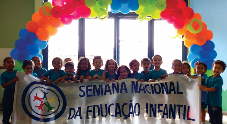 Cerca de 16 crianças seguram a faixa com os dizeres: "Semana nacional da educação infantil" tendo atrás uma guirlanda de balões. 