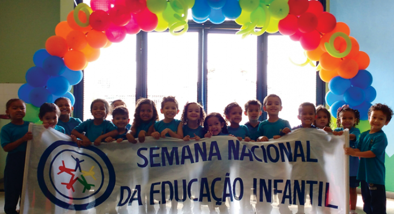 Formação para professores, oficinas, visitas a espaços culturais, excursões e passeata fazem parte da Semana Nacional da Educação Infantil. Foto: Smed/PBH Cerca de 16 crianças seguram a faixa com os dizeres: "Semana nacional da educação infantil" tendo atrás uma guirlanda de balões.