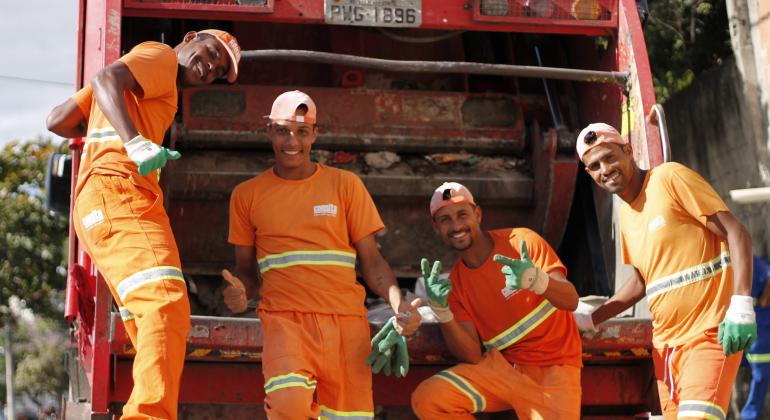 Profissionais relatam as alegrias e os desafios de um cotidiano inusitado pelas ruas e avenidas de Belo Horizonte. Foto: Ana Clara Nunes/PBH Quatro garis da SLU, com uniformes laranjas, atrás do caminhão de lixo.