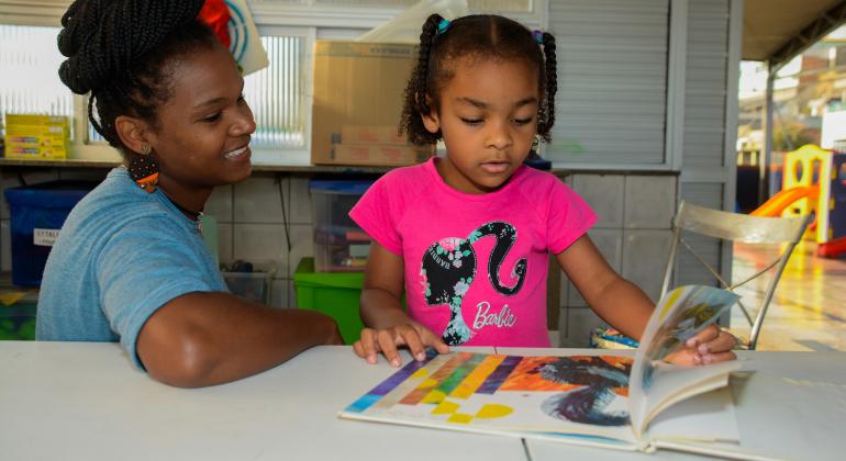 Projeto literário municipal visa a estimular o hábito da leitura nas crianças desde a primeira infância. Foto: Stênio Lima/PBH Mulher acompanha criança na leitura de livro infantil.