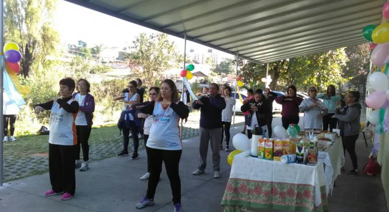 Usuários do Centro de Saúde Califórnia, na região Noroeste, celebram 4 anos de prática do Programa Lian Gong. Foto: Divulgação Cerca de 10 usuários fazem Liang Gong em área coberta; ao lado, mesa com refrigerantes e quitutes.