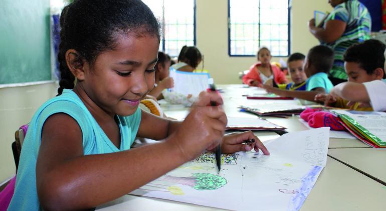Aprovados começarão a ser convocados para atuarem na Rede Municipal de Ensino neste mês. Foto: Cláudio Lacerda Criança morena de camiseta verde, sentada à mesa, faz um desenho no papel ofício.