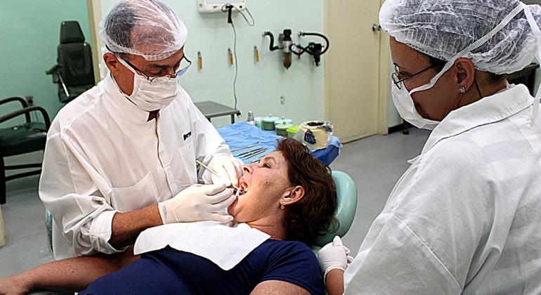 Dentista atende paciente acompanhado de auxiliar. Foto ilustrativa.