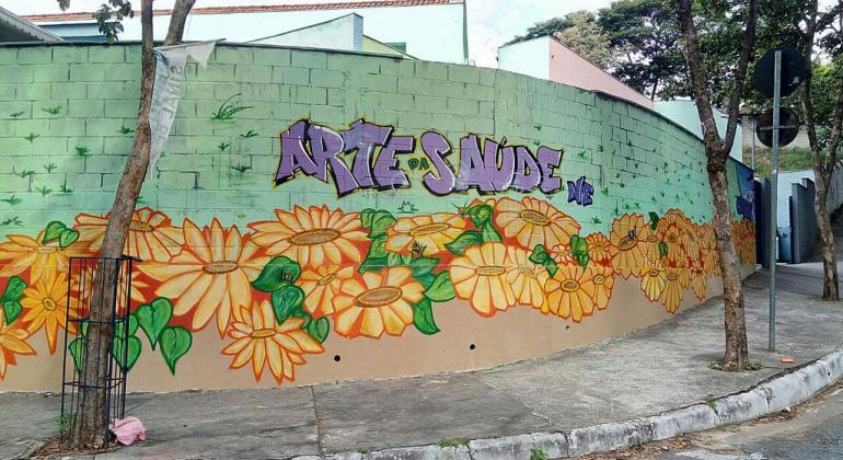 Muro de uma esquina pintado de verde com flores de pétalas amarelas e os dizeres "Arte da Saúde" em roxo.