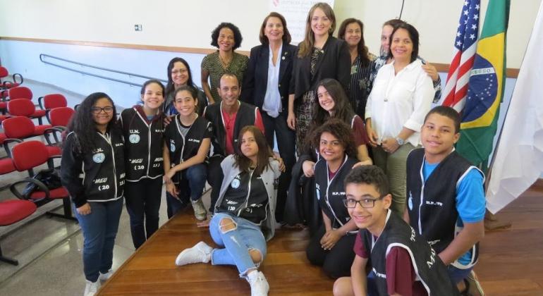 Cerca de 14l pessoas, entre alunos e professores, em cima de um palco com as bandeiras do Brasil e dos Estados Unidos ao fundo.