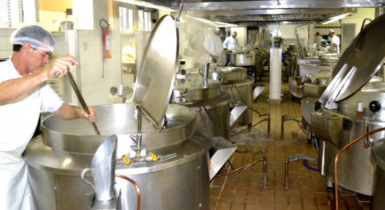 Cozinheiro prepara almoço em panela muito grande de retaurante popular. Ao lado e ao fundo, outras quatro panelas do mesmo tamanho e dois cozinheiros.