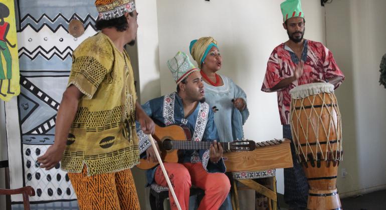 Três homens e uma mulher trajados com roupa africana em um palco. Um toca um violão, outro toca um tambor e a mulher toca um xilofone. O outro homem dança.